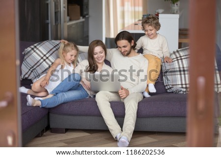 Married couple and little children sitting on couch in living room. Parents with little daughter, son looking at computer and smiling. Happy young family together at home watching funny videos online