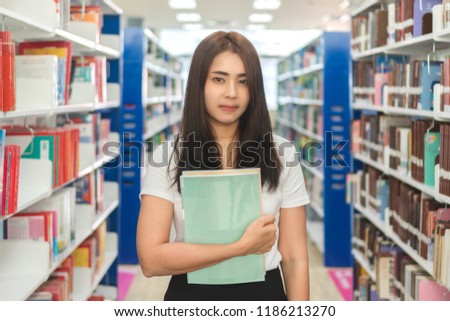 Portrait of young asian female college with book standing in library. Beautiful teenager student holding book and smiling to camera at university. Education, learning and research concept.
