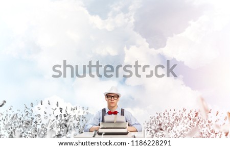 Young man writer in hat and eyeglasses using typing machine while sitting at the table among flying letters with cloudy skyscape on background.