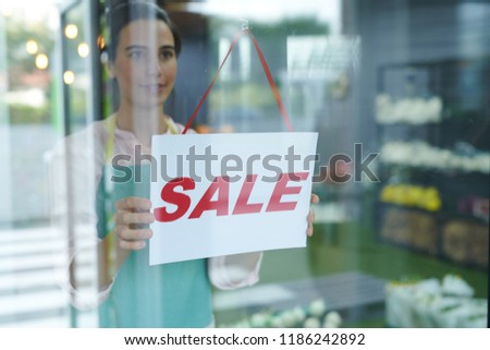 Waist up portrait of cheerful female shopkeeper hanging red SALE sign on glass door or window