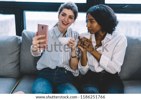 Positive multicultural female bloggers with coffee in hands positing at front camera and making funny selfie on modern smartphone.Cheerful young women resting at home and taking photo on cellular