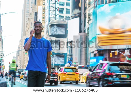 Young African American Man traveling in New York, wearing blue T shirt, walking on busy street in Times Square of Manhattan, talking on cell phone. High buildings, billboards, cars, bike on background