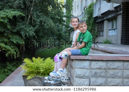 Teenager girl and boy in roller skates with protection on knees sitting on stairs with blurred building on background 