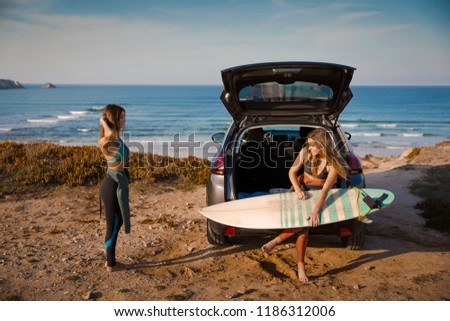 Two beautiful surfer girls near the coastline with her car, and getting ready for surfing