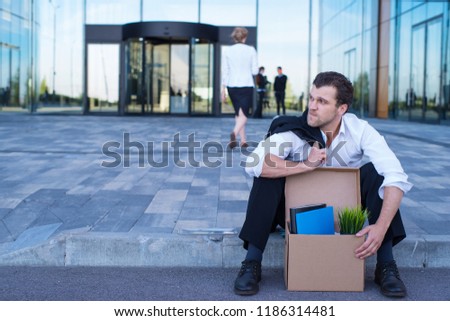Fired business man sitting frustrated and upset on the street near office building with box of his belongings. He lost work Royalty-Free Stock Photo #1186314481