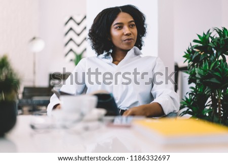Charming African American young woman in casual wear looking away while writing blog article at home. Attractive dark skinned hipster girl with fashion magazine in hands relaxing in modern apartment
