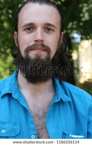 Hairy young man, with a beard, wearing an unbuttoned blue shirt against the background of green trees