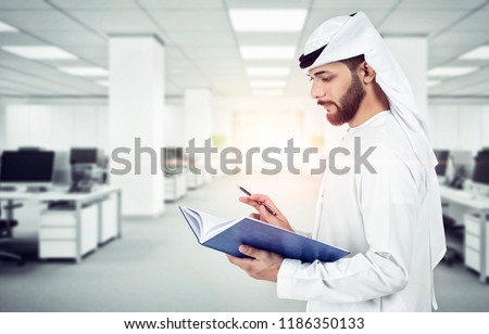 Arab Emirati man writing on a book, working in office  