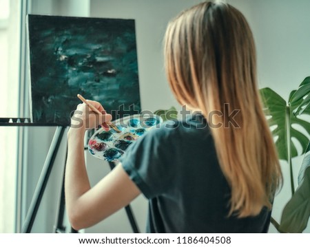 Cute beautiful girl artist painting a picture on a canvas on an easel. Space for text. Long hair. Holding colorful brush and palette.