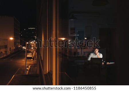 Young Asian businessman sitting at his desk working on a laptop late at night behind office building windows in the city at night Royalty-Free Stock Photo #1186450855