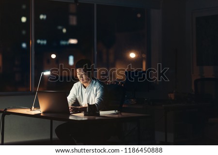 Young Asian businessman working on a laptop while sitting at his desk in a dark office at night with city lights in the background Royalty-Free Stock Photo #1186450888