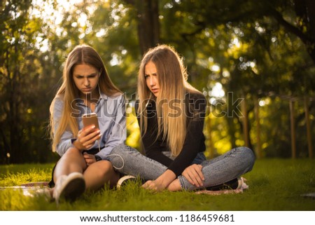 Photo of confused displeased girls sisters students sitting in the park outdoors on grass using mobile phone listening music with earphones.