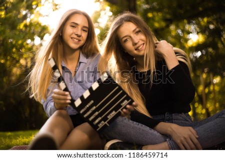 Photo of beautiful happy girls sisters students sitting in the park outdoors on grass have a rest holding film making clapperboard.