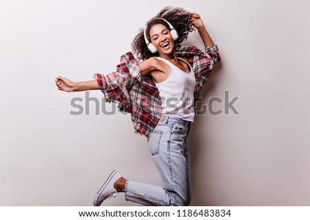 Cute curly girl in jeans jumping with happy face expression. Good-humoured african lady in headphones expressing positive emotions during photoshoot on white background.