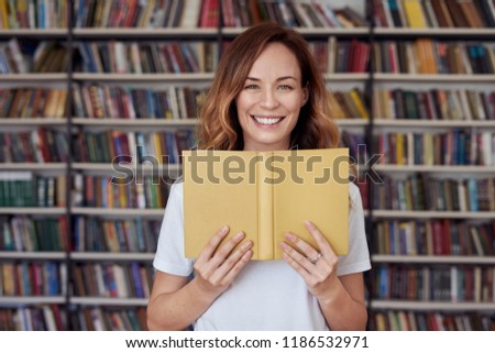 Portrait of smiling woman model with opened book in a library, bookshelf behind, long hair. Hipster college student lady.
