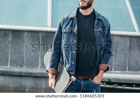 Young serious guy in black T-shirt looking in camera holding skateboard in hand with modern background