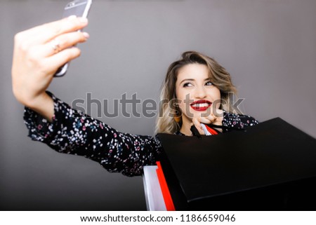 Young woman with red lips and fair hair in patterned blouse making selfie with shopping bags on smartphone on gray background