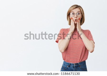 Wow, favotire author released new book. Impressed and surprised good-looking female in glasses with short haircut, folding lips and gasping, holding palms on cheeks, being amazed and excited Royalty-Free Stock Photo #1186671130