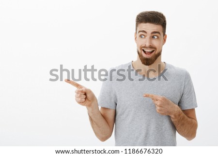 Enthusiastic guy feeling happy and amazed gazing at super cool copy space. Portrait of handsome delighted and charismatic man with beard and moustache looking and pointing left over gray wall