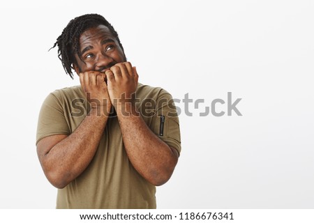 Studio shot of cute impatient dark-skinned man in olive t-shirt, holding hands near jaw, biting fingernails and gazing with temptation at upper right corner, waiting for surprise with excitement