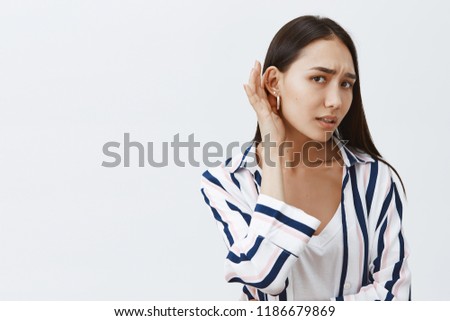 Studio shot of good-looking gloomy girlfriend with long dark hair, leaning towards camera with one ear and gazing at camera, eavesdropping and whispering, hearing rumor, feeling nervous Royalty-Free Stock Photo #1186679869