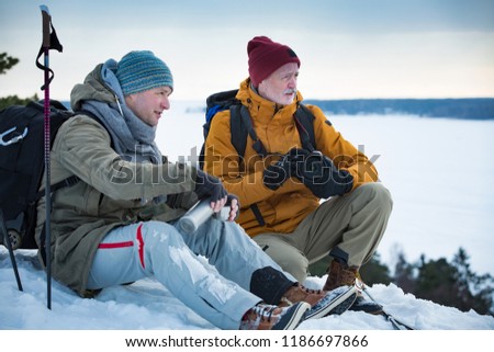 Two mature men exploring Finland in winter. Hikers sit on top of rock, take pictures with camera, drink hot coffee from thermos flask. Northern landscape with frozen Baltic Sea and snowy islands.