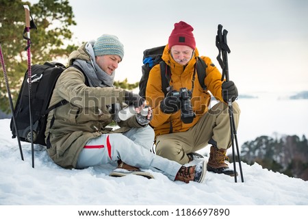 Two mature men exploring Finland in winter. Hikers sit on top of rock, take pictures with camera, drink hot coffee from thermos flask. Northern landscape with frozen Baltic Sea and snowy islands.