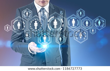 Unrecognizable young businessman looking at his smartphone screen with people network holgoram standing against blue background. HR concept. Toned image double exposure