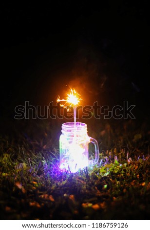 Festive card with bright colorful garland lights and Bengali fire or sparkler in glass jar on night nature background.