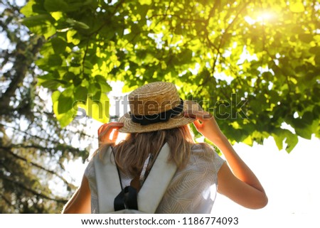 Stylish hipster girl with a straw hat  is standing outdoors. Woman in blue dress. Silver grey backpack. Travel holiday concept. Summer lifestyle photo. Shades of sun and trees.