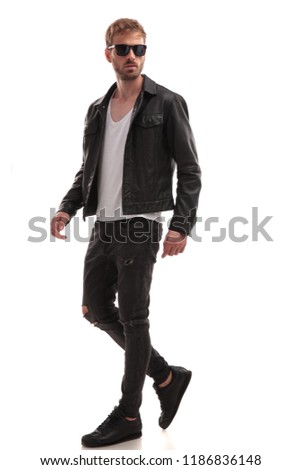 side view of cool young man with sunglasses and black leather jacket walking on white background, full length picture