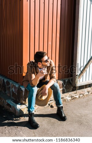 handsome cowboy in sunglasses sitting and smoking cigarette at ranch