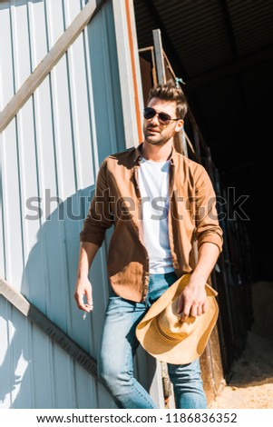 handsome man in sunglasses holding cigarette and hat, leaning on wall at ranch