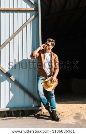 handsome man in sunglasses smoking cigarette, holding hat and leaning on wall at ranch