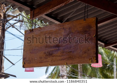 Blank wooden board sign hang under roof. Tropical island cafe or hotel sign mockup. Wooden board with palm leaf background. Exotic place for vacation. Empty billboard outdoor. Rustic board texture