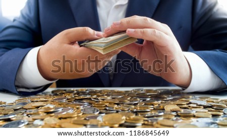 Closeup photo of young successful businessman sitting behind table covered with golden coins and holding stack of money