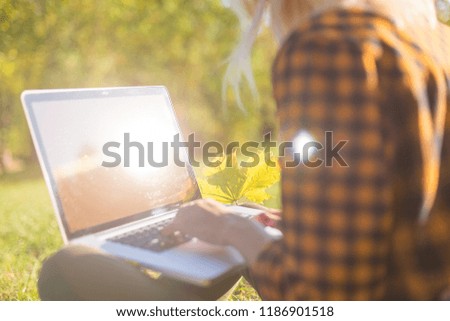 Girl are typing on a laptop keyboard on a autumn background with fallen leaves and green grass.  Online work, freelancing. Side top View. Sunlight on the screen

