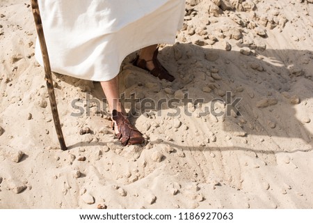 cropped image of Jesus in robe and sandals walking on sand with wooden staff in desert Royalty-Free Stock Photo #1186927063