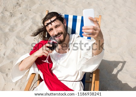 smiling Jesus resting on sun lounger, drinking wine and taking selfie with smartphone in desert