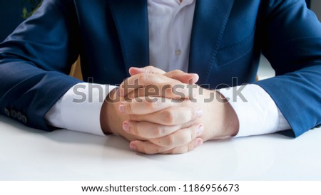 Closeup photo of young businessman in blue suit sitting behind white wooden desk at office with folded hands