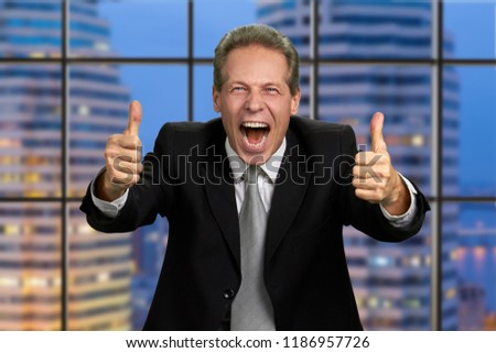 Excited businessman expresses hapiness. Ecstatic caucasian businessman expresses satisfaction and triumph with thumbs up on skyscraper background.