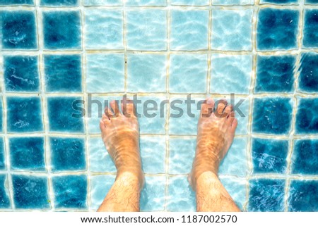 Top view selfie the feet of an Asian man feet standing in the pool. Feet and Clear blue water with pattern on the edge of the pool with small square grid pattern.