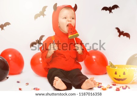 Tricky cute baby boy in costume devil sitting on a background of decorations for Halloween and eating red watermelon lollipop.