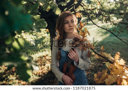 Beautiful young woman playing Ukulele feeling carefree and happy in the natural autumn sunlight warm tone in a park, creative activity.