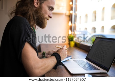 Serious bearded Sweden man with long hair having online training course via laptop computer, writing notary while sitting in modern coworking space. Male project manager working distance on netbook 