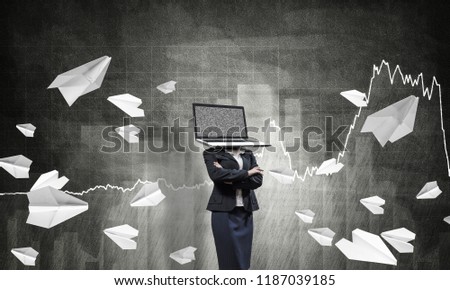 Business woman in suit with laptop instead of head keeping arms crossed while standing against flying paper planes and analytical charts drawn on dark wall on background.