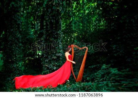 Portrait of a woman with a harp.Beautiful brown-haired woman with a flower wreath on her head,
 wearing a red long dress playing the harp in the forest. Royalty-Free Stock Photo #1187063896