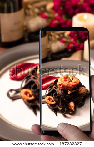 Photographing with smartphone in hand. food concept with Black spaghetti. Black seafood pasta with shrimp in the white plate and red rose and grape wine. Mediterranean delicacy food. selective focus/s