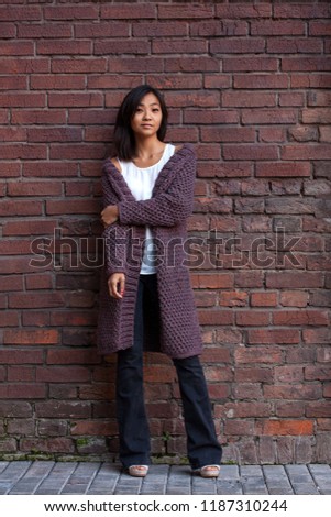 beautiful Asian young woman standing in a knitted purple warm cardigan, hugging herself over the shoulder with one hand standing against a brick wall