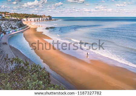 Low tide at Stone Bay, Broadstairs, Kent as summer turns to autumn, a lone surfer walks on the beach and a family on the promenade along side the beach huts and white cliffs. Royalty-Free Stock Photo #1187327782
